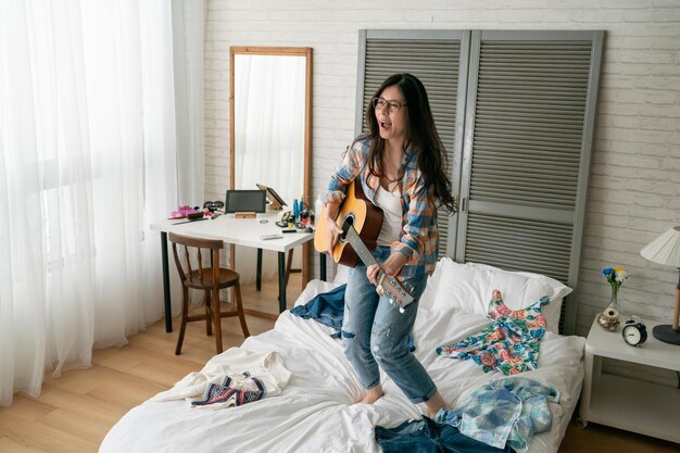 retrato de una joven china asiática feliz tocando la guitarra y bailando en la cama. mujer loca divirtiéndose saltando en el dormitorio. estudiante universitaria ama la música disfruta unirse a la práctica de la banda en casa.