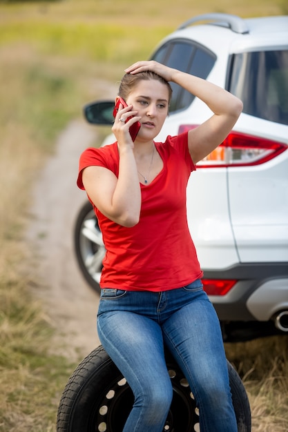 Retrato de joven conductora sentada junto al coche roto y hablando por teléfono