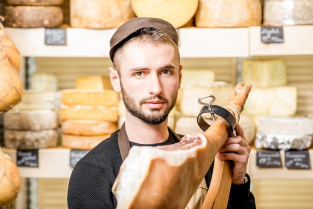 Retrato de un joven contador con pierna de jamón de pie delante del escaparate de la tienda lleno de quesos