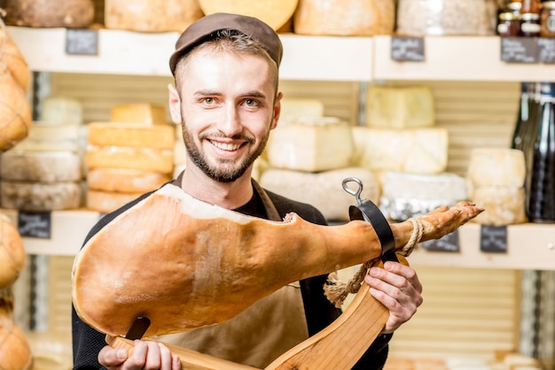 Retrato de un joven contador con pierna de jamón de pie delante del escaparate de la tienda lleno de quesos