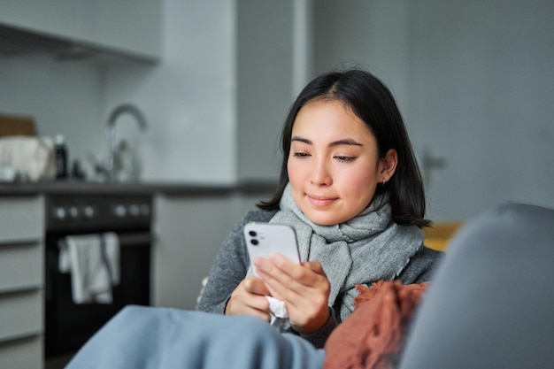 Retrato de una joven coreana enferma sentada en un sofá enviando un mensaje de texto usando un teléfono móvil para contactarla