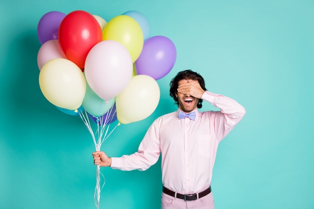 Retrato de joven cubren los ojos vestidos con ropa formal mantenga globos de colores fiesta sorpresa aislado de fondo de color verde azulado