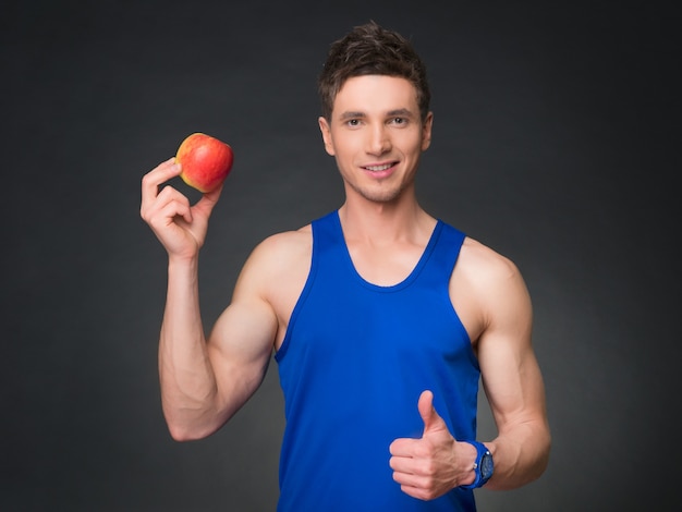 Retrato de joven culturista sonriente sosteniendo manzana en fondo negro.