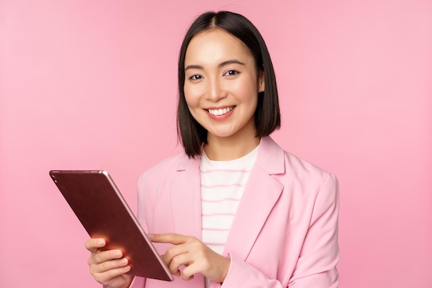 Retrato de una joven dama de oficina corporativa asiática con tableta digital con traje sonriendo y luciendo profesional posando sobre fondo rosa