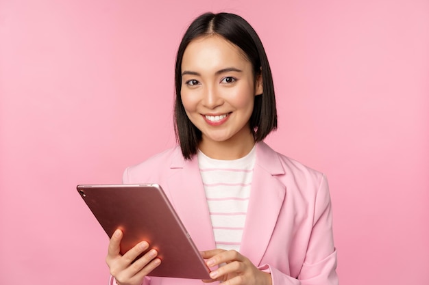 Retrato de una joven dama de oficina corporativa asiática con tableta digital con traje sonriendo y luciendo profesional posando sobre fondo rosa