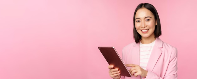 Retrato de una joven dama de oficina corporativa asiática con tableta digital con traje sonriendo y luciendo profesional posando sobre fondo rosa