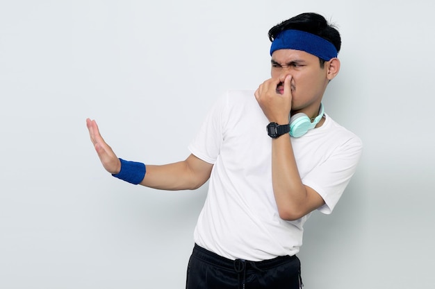 Retrato de un joven deportista asiático serio con diadema azul y camiseta blanca con auriculares hace un gesto de parada y se cubre la nariz con la mano aislada sobre fondo blanco Concepto de deporte de entrenamiento