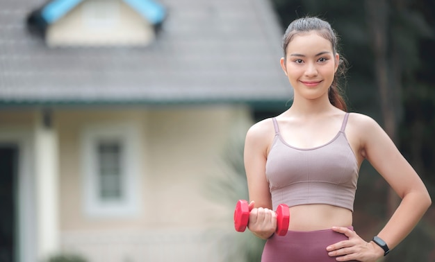 Retrato de joven deportista hermosa sosteniendo mancuernas rojas, sonriendo y mirando a la cámara mientras está de pie al aire libre frente a la casa.