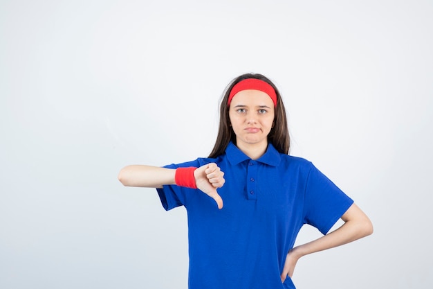 Retrato de una joven deportista mostrando el pulgar hacia abajo sobre una pared blanca