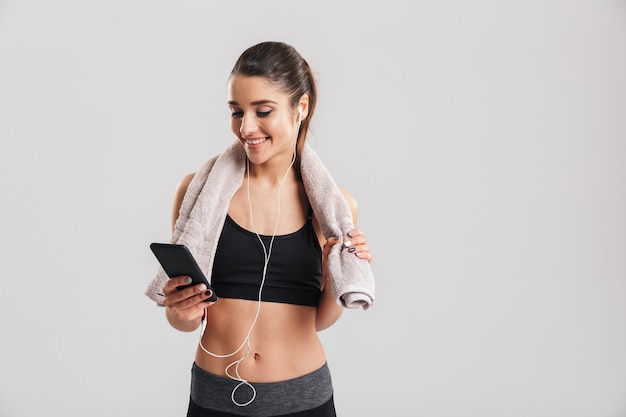 Retrato de joven deportiva en chándal posando con una toalla en el cuello y escuchando música a través de auriculares, aislado sobre la pared gris