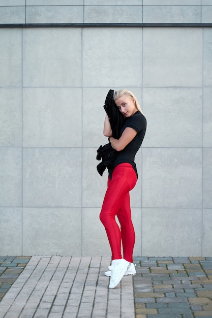 Retrato de joven deportiva positiva vistiendo ropa deportiva negra, polainas rojas y zapatillas blancas de moda. Tiro al aire libre en la pared gris