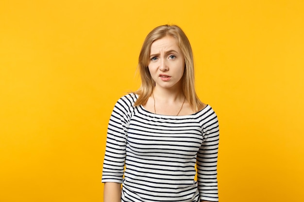 Retrato de una joven desconcertada y descontenta con ropa a rayas de pie, mirando una cámara aislada en un fondo amarillo anaranjado en el estudio. Emociones sinceras de la gente, concepto de estilo de vida. Simulacros de espacio de copia.