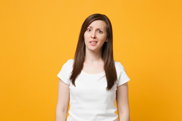 Retrato de una joven desconcertada perpleja con ropa informal blanca de pie y mirando una cámara aislada en un fondo de pared naranja amarillo brillante en el estudio. Concepto de estilo de vida de las personas. Simulacros de espacio de copia.