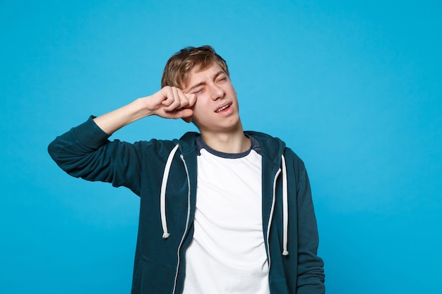 Retrato de joven descontento insatisfecho en ropa casual llorando y secándose las lágrimas aisladas en la pared azul. Personas sinceras emociones, concepto de estilo de vida.