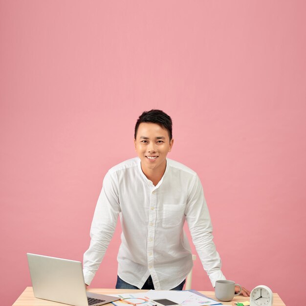 Retrato de un joven diseñador asiático sonriente que trabaja en una laptop mientras se apoya en su escritorio