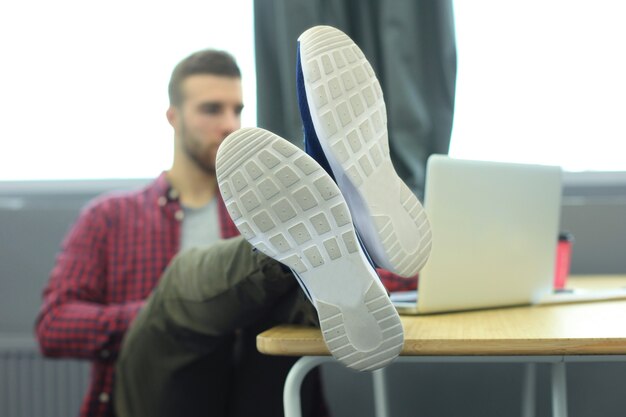 Retrato de un joven diseñador muy relajado que se inclina hacia atrás en su escritorio y pone los pies sobre su escritorio en la oficina.