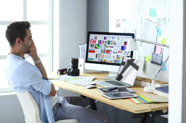 Retrato de un joven diseñador sentado en un estudio gráfico frente a una laptop y una computadora mientras trabaja en línea