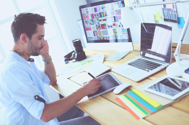 Retrato de un joven diseñador sentado en un estudio gráfico frente a una laptop y una computadora mientras trabaja en línea