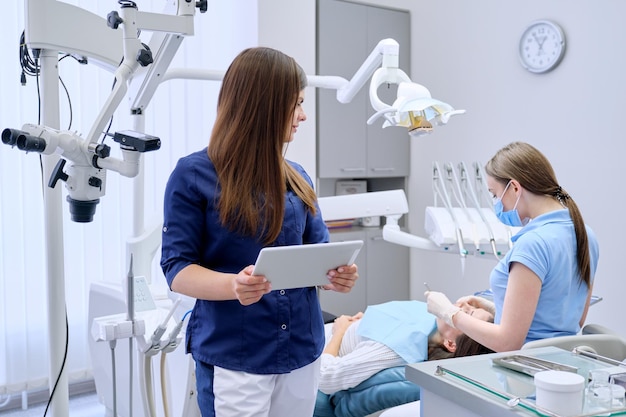 Retrato de una joven doctora dentista con tableta digital en el consultorio dental, tratando a una paciente en el fondo de una silla. Concepto de medicina, odontología y atención de la salud.