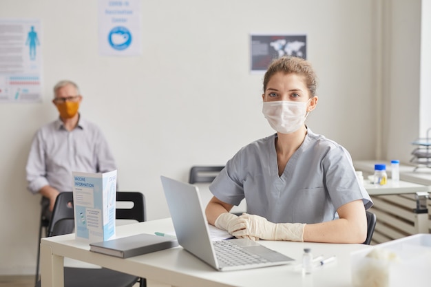 Retrato de joven doctora con máscara y mirando a la cámara mientras espera a los pacientes en la clínica o centro de vacunación, espacio de copia