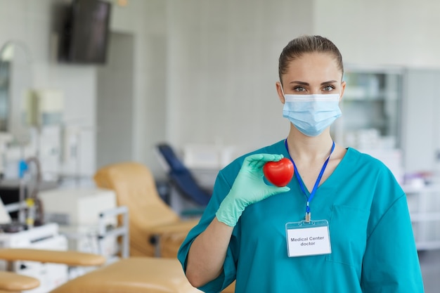 Retrato de joven doctora en máscara y uniforme con forma de corazón en sus manos y