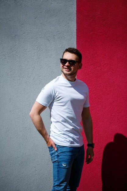 Retrato de un joven elegante, un hombre vestido con una camiseta blanca en blanco de pie sobre un fondo de pared gris y rojo. Estilo urbano de ropa, imagen de moda moderna. Moda de hombres