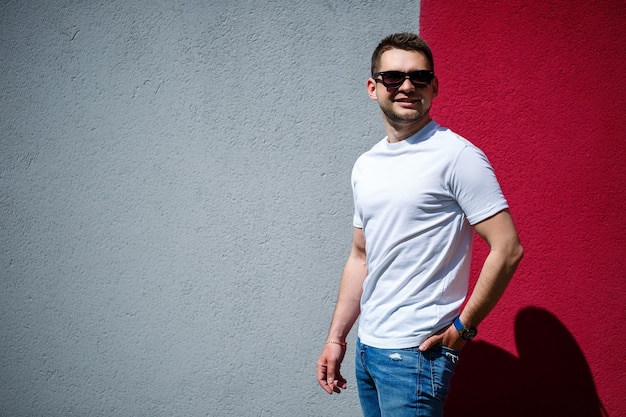 Retrato de un joven elegante, un hombre vestido con una camiseta blanca en blanco de pie sobre un fondo de pared gris y rojo. Estilo urbano de ropa, imagen de moda moderna. Moda de hombres