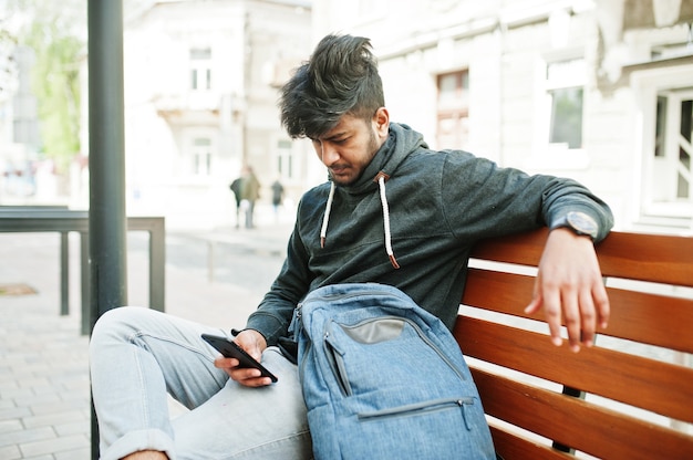 Retrato de joven elegante modelo de hombre indio posan en la calle, sentado en un banco y sostienen el teléfono inteligente en la mano.