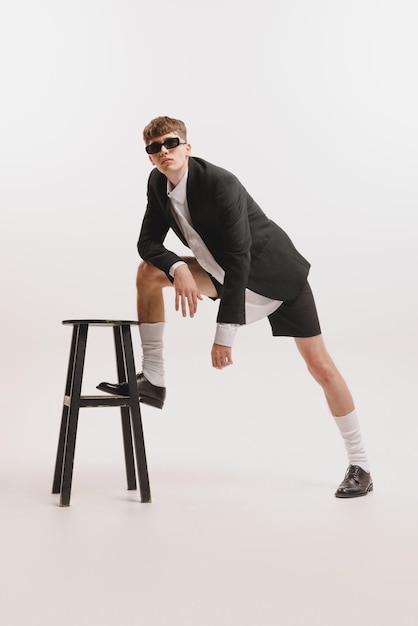 Retrato de un joven elegante posando con un traje de moda apoyado en una silla aislada sobre un fondo blanco de estudio