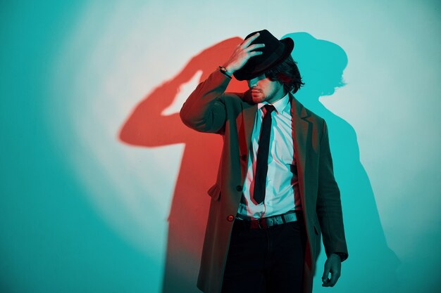 Retrato de joven elegante con sombrero, traje y corbata que se encuentra en luces de neón en el estudio.