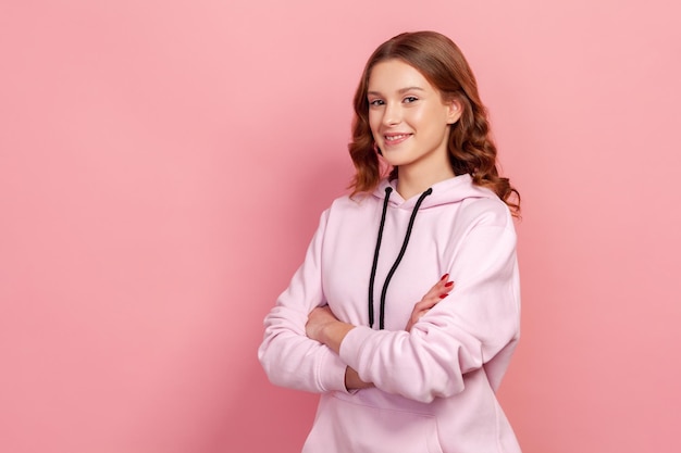 Retrato de una joven emocionada de cabello rizado con capucha de pie con las manos cruzadas y una sonrisa en un estudio interior aislado en un fondo rosa