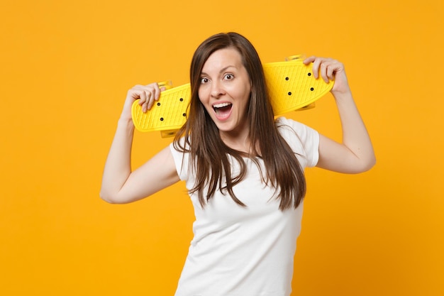 Retrato de una joven emocionada con ropa informal blanca que mantiene la boca abierta, sostiene una patineta amarilla aislada en un fondo naranja amarillo brillante en el estudio. Concepto de estilo de vida de las personas. Simulacros de espacio de copia.