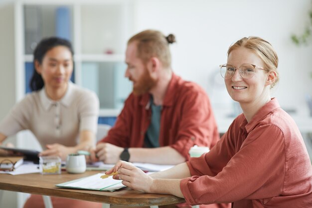 Retrato de joven empresaria en anteojos sonriendo mientras está sentado en la mesa durante la reunión de negocios en la oficina