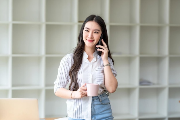 Retrato de una joven empresaria asiática sonriente parada sosteniendo una taza de café y hablando por teléfono móvil en la oficina.