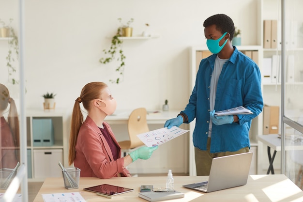 Retrato de joven empresaria con máscara y guantes entregando documentos a un colega afroamericano mientras trabajaba en la oficina posterior a la pandemia