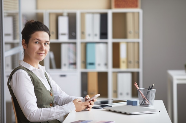 Retrato de joven empresaria mirando al frente mientras está sentado en el lugar de trabajo y usa su teléfono móvil en la oficina