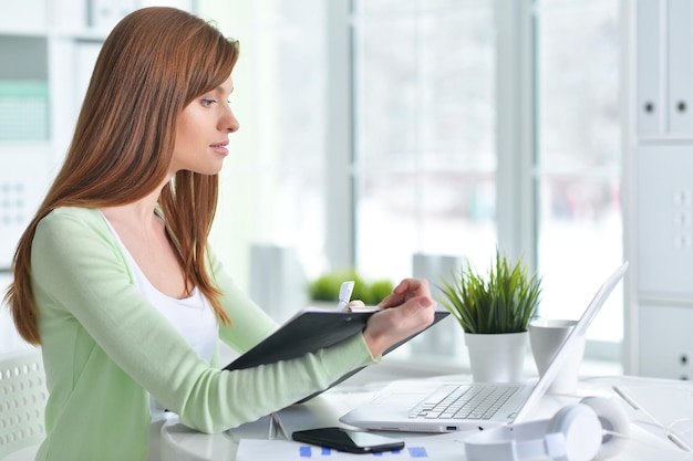 Retrato de una joven empresaria mirando la pantalla de una laptop y escribiendo información en el portapapeles