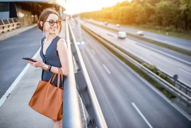 Retrato de una joven empresaria de pie con el teléfono en la carretera durante el viaje de negocios al atardecer