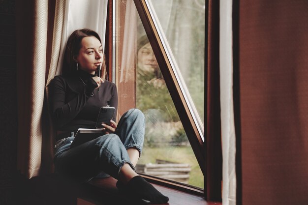 Retrato de joven empresaria con portátil y teléfono en la ventana del salón en casa de campo. Mujer adicta al trabajo en ropa casual de casa trabajando en vacaciones. Inspiración creativa y negocios
