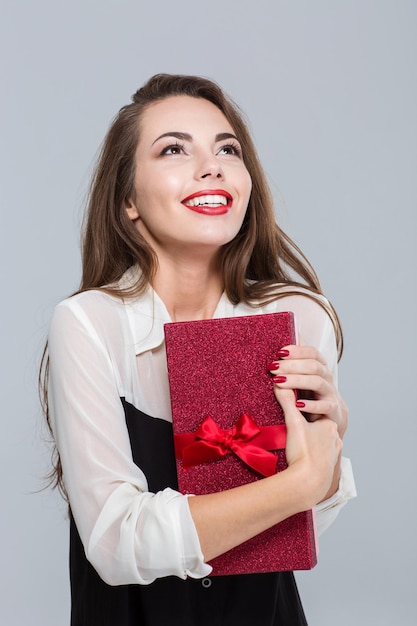 Retrato de una joven empresaria sonriente sosteniendo una caja de regalo y mirando hacia arriba sobre la pared gris