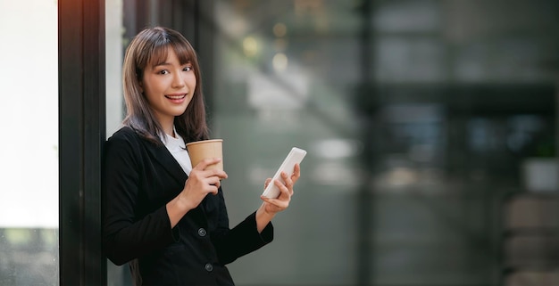 Retrato de una joven empresaria sosteniendo una taza de café y usando un teléfono móvil de pie en la oficina sonriendo y mirando la cámara