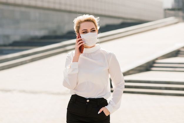 Retrato de una joven empresaria con teléfono, portátil, tableta, café al aire libre. Bonita chica rubia en guantes de goma azul y máscara.