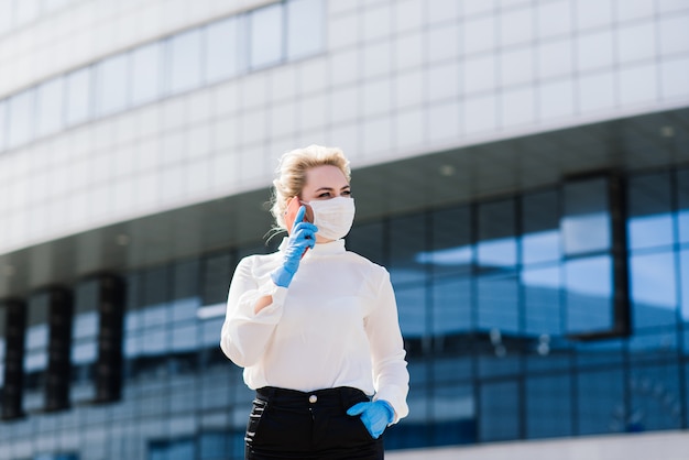 Foto retrato de una joven empresaria con teléfono, portátil, tableta, café al aire libre. chica rubia en guantes de goma azul y máscara.