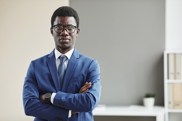 Retrato de joven empresario africano en anteojos y en ropa formal de pie con los brazos cruzados.