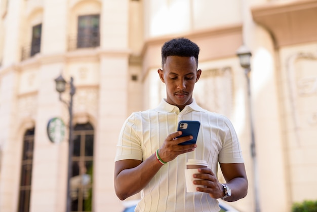 Retrato de joven empresario africano con ropa casual mientras envía mensajes de texto con el teléfono móvil y sostiene la taza de café para llevar