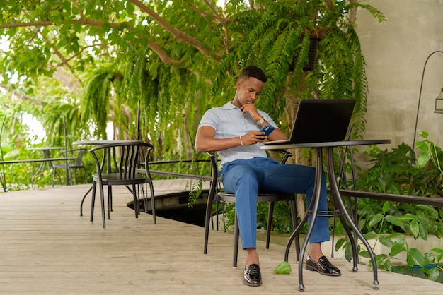 Retrato de joven empresario africano vistiendo ropa casual y sentado en la cafetería mientras usa una computadora portátil y un teléfono mientras que el distanciamiento social