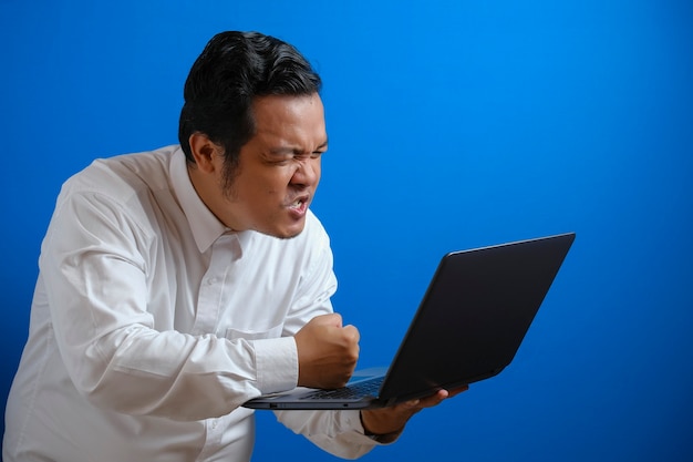 Retrato de joven empresario asiático escribiendo en el portátil, expresión enojada loca, sobre fondo de pared azul con espacio de copia