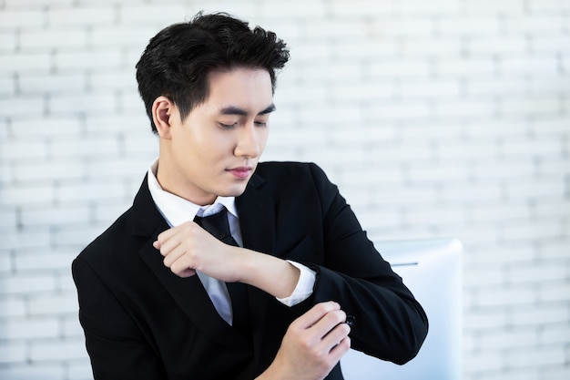 Retrato del joven empresario asiático de humor feliz pone gemelos con un traje de hombre con chaqueta negra y camisa blanca en la oficina de la pared blanca