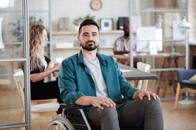 Retrato de joven empresario barbudo sentado en silla de ruedas y mirando a cámara mientras trabaja en la oficina