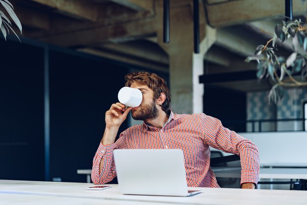 Retrato de joven empresario caucásico usando la computadora portátil en su lugar de trabajo en la oficina moderna